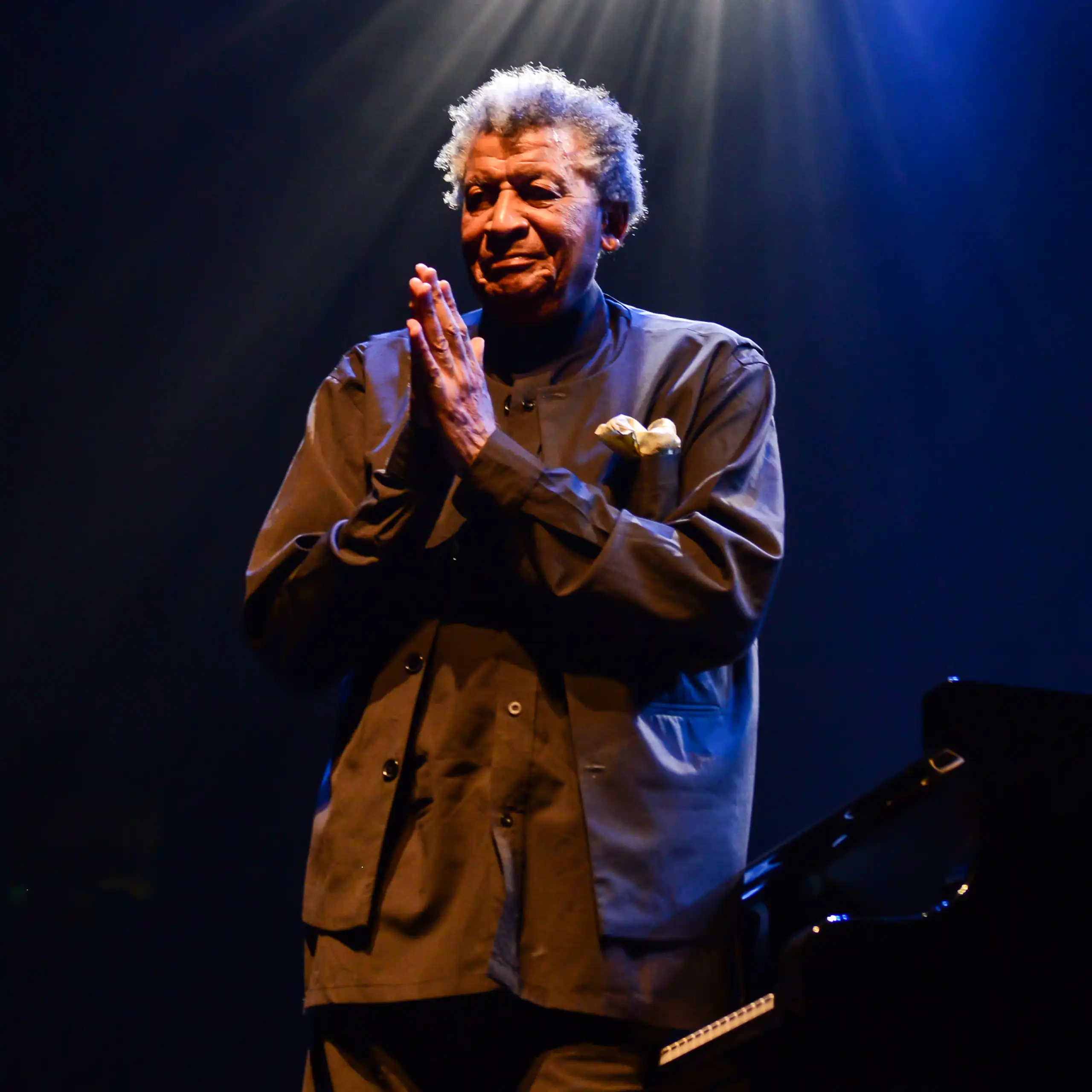 An elderly man with grey hair stands under a light on a stage next to a piano, hands in a thank-you prayer position in front of him.