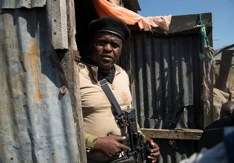 A man wearing a beige t-shirt and wearing a hat, with an assault rifle in his hands.
