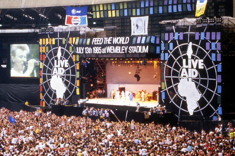 A huge outdoor concert with thousands of people cheering for David Bowie onstage.