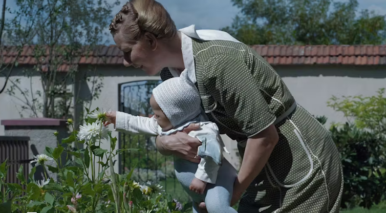 A woman with a baby bending over a flower in a sunny garden.