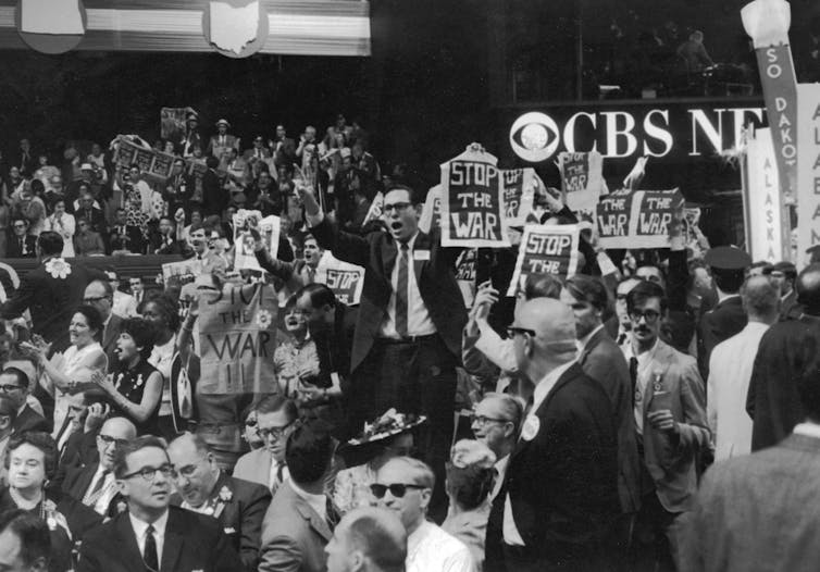 A crowd of people in a convention hall, with some holding signs that say 'Stop the war.'
