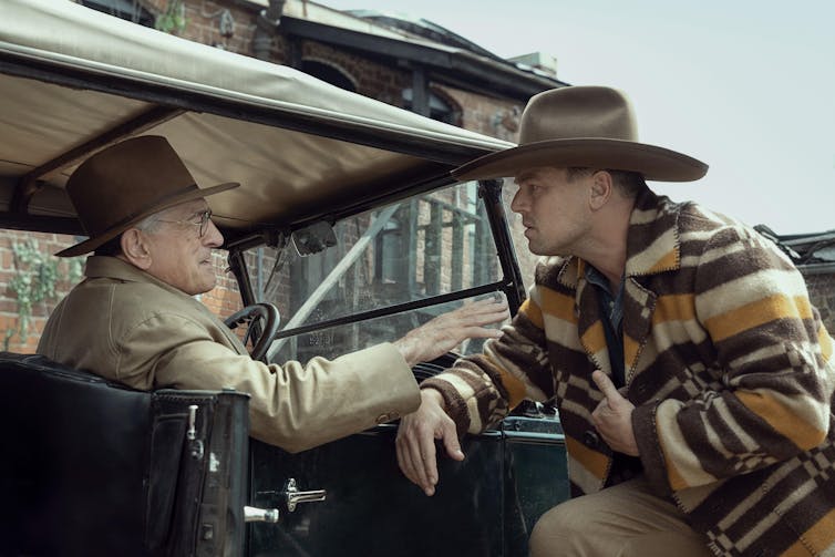Two early 20th-century men in cowboy hats talking in a car.