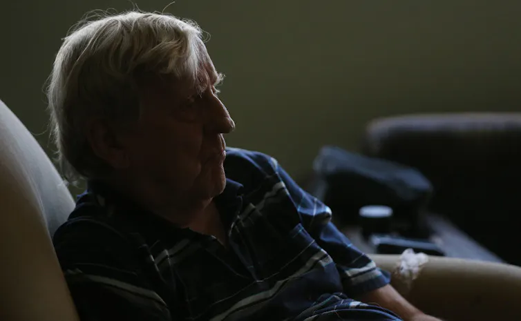 A man with white hair, wearing a blue striped shirt, sits somberly in a shadowed room.