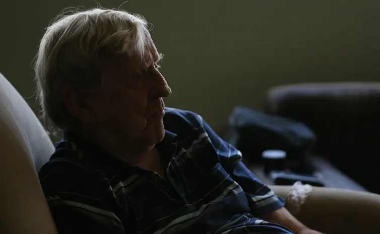A man with white hair, wearing a blue striped shirt, sits somberly in a shadowed room.