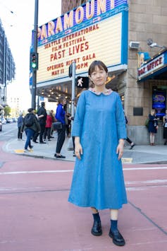Asian woman in a blue dress stands on a street in front of a big, bright billboard advertising a screening for 'Past Lives.'
