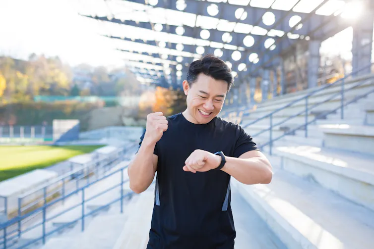 A man in a black T-shirt near a sports field looking at his smartwatch and smiling