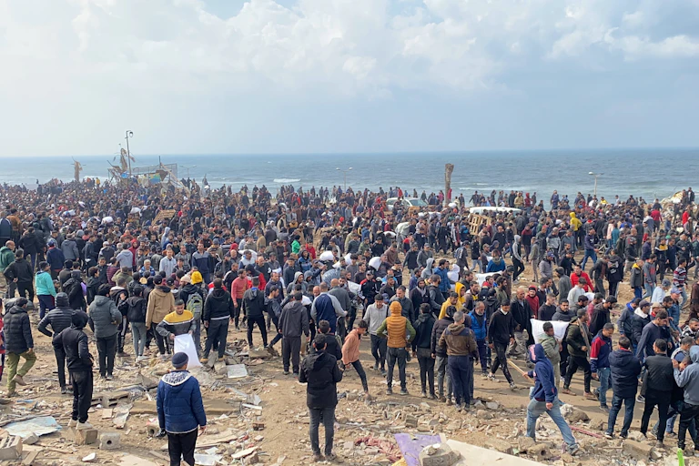 Hundreds of people gathered at a beach, which has rubble scattered around.