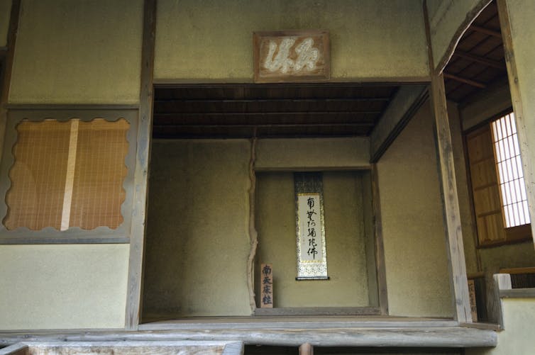 The interior of a simple room with faded walls, wooden beams, and a simple scroll hanging in the background.