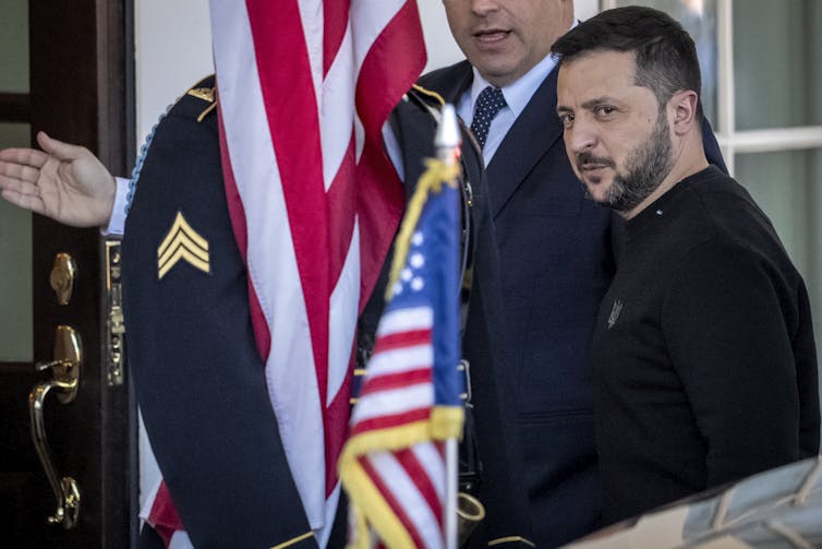 A man in a dark sweater and beard enters a building. U.S. flags are seen around him.