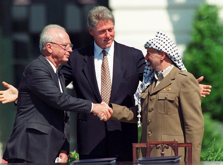 Three men two in suits one wearing a traditional Palestinian headscarf stand. Two shake hands.