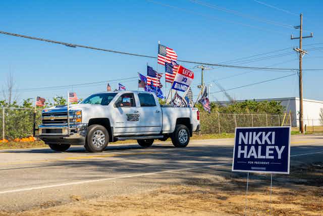A truck festooned with American flags and Trump signs drives past a Nikki Haley campaign sign.