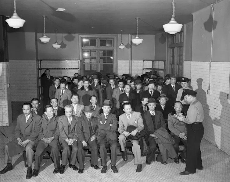 A group of men wearing suits and hats sit while a uniformed man stands beside them.