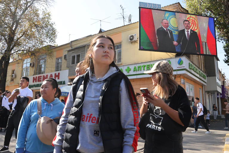 Women stand in a group in front of shops and a big TV screen showing two men.