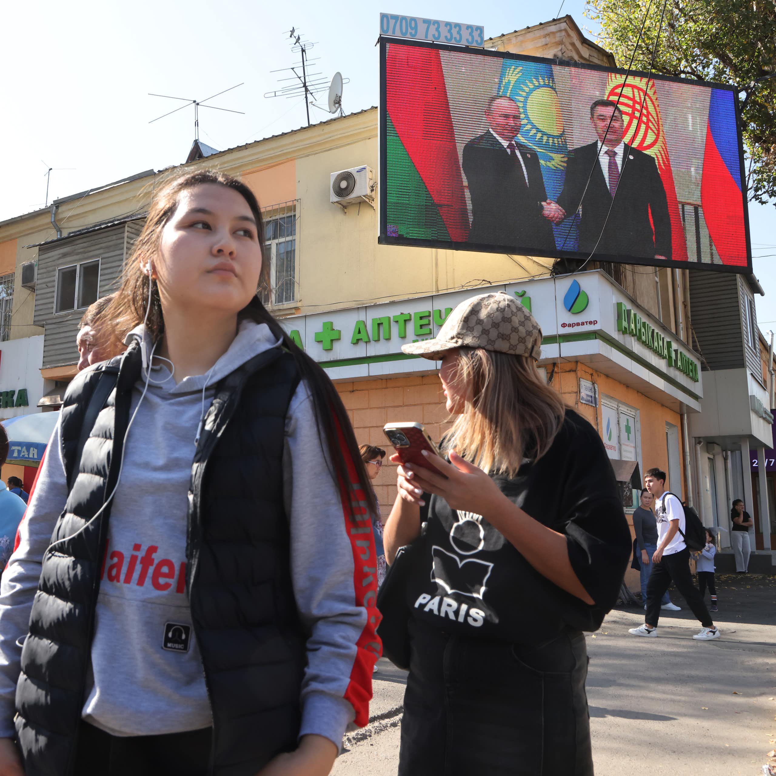 Women stand in a group in front of shops and a big TV screen showing two men.