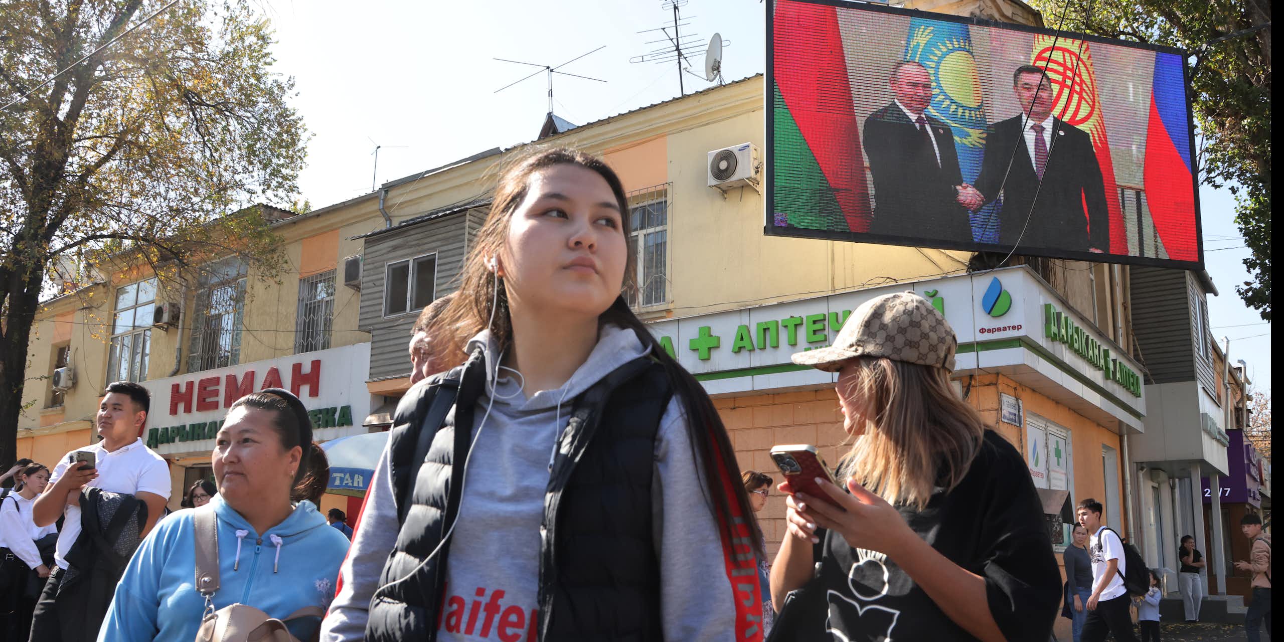 Women stand in a group in front of shops and a big TV screen showing two men.