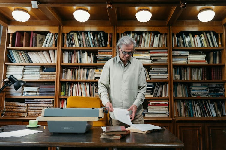 An older man with solver hair sorts through papers and books in a study.