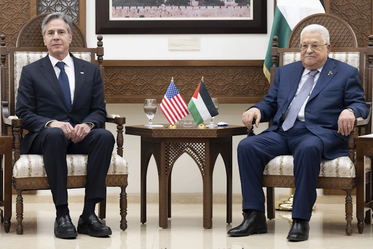 Two men sit with a table between them with Palestinian and US flags on.