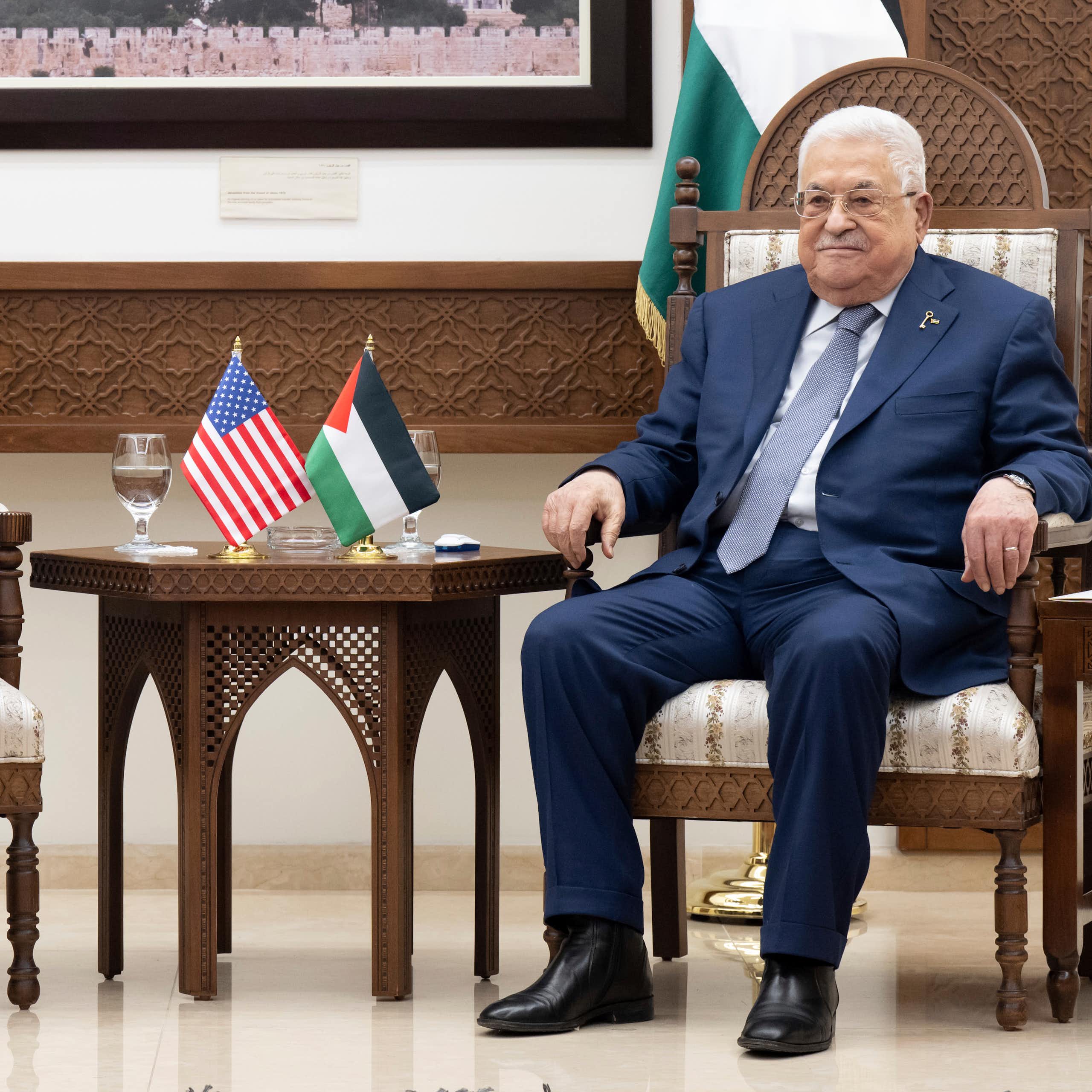 Two men sit with a table between them with Palestinian and US flags on.