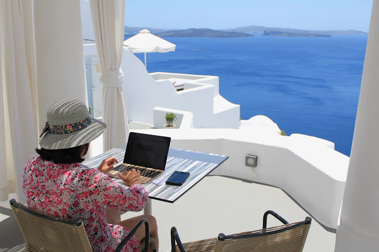 A woman works in her laptop by a window overlooking a Greek village.
