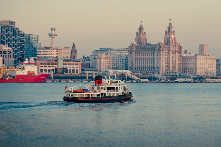 Mersey ferry boat moving across on river, urban Liverpool skyline in background with lots of distinctive landmark buildings, grey sky