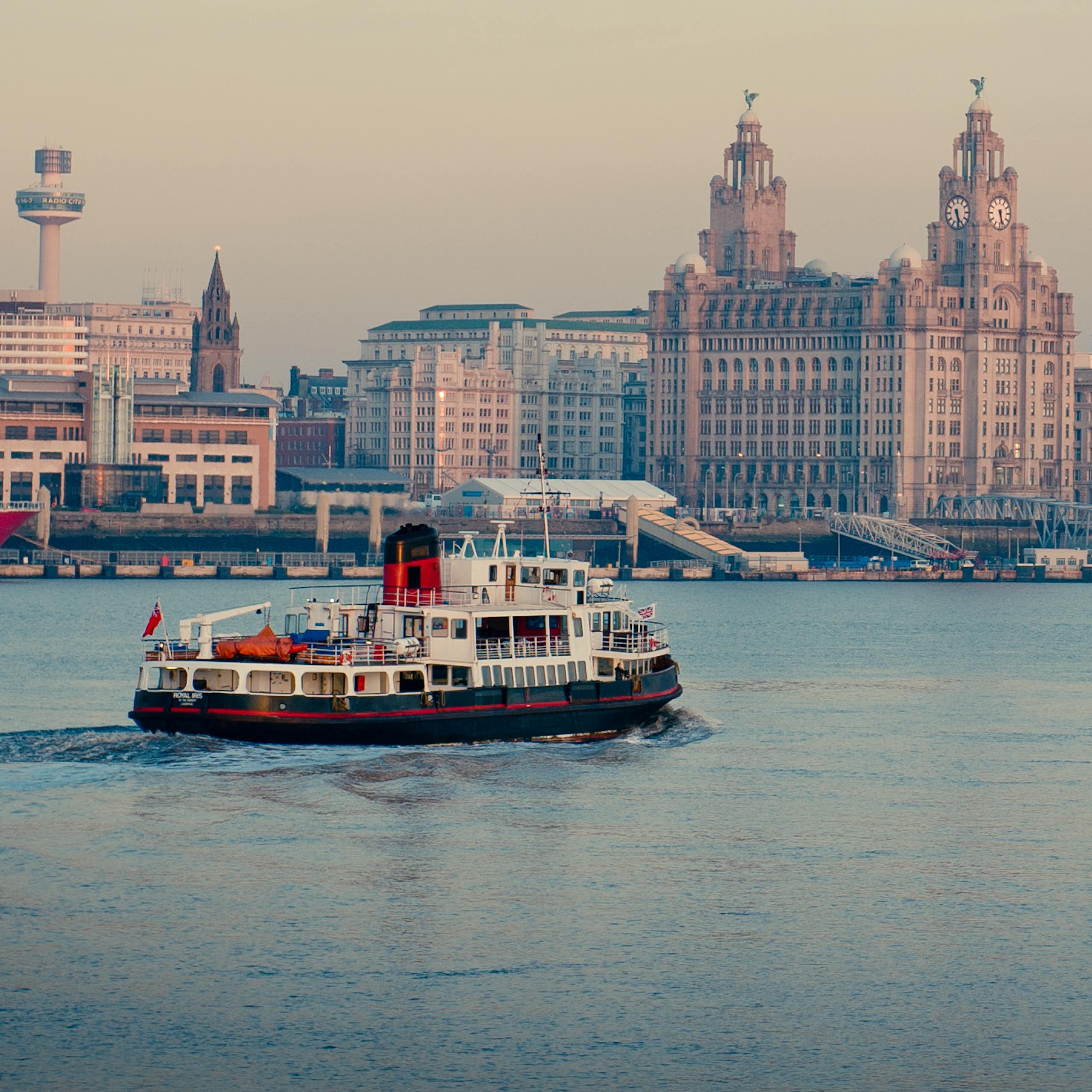 Mersey ferry boat moving across on river, urban Liverpool skyline in background with lots of distinctive landmark buildings, grey sky
