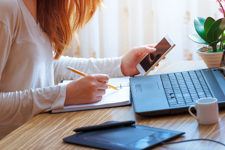 Une jeune femme assise à un bureau, devant un ordinateur portable, consulte son téléphone et écrit en même-temps dans un carnet.