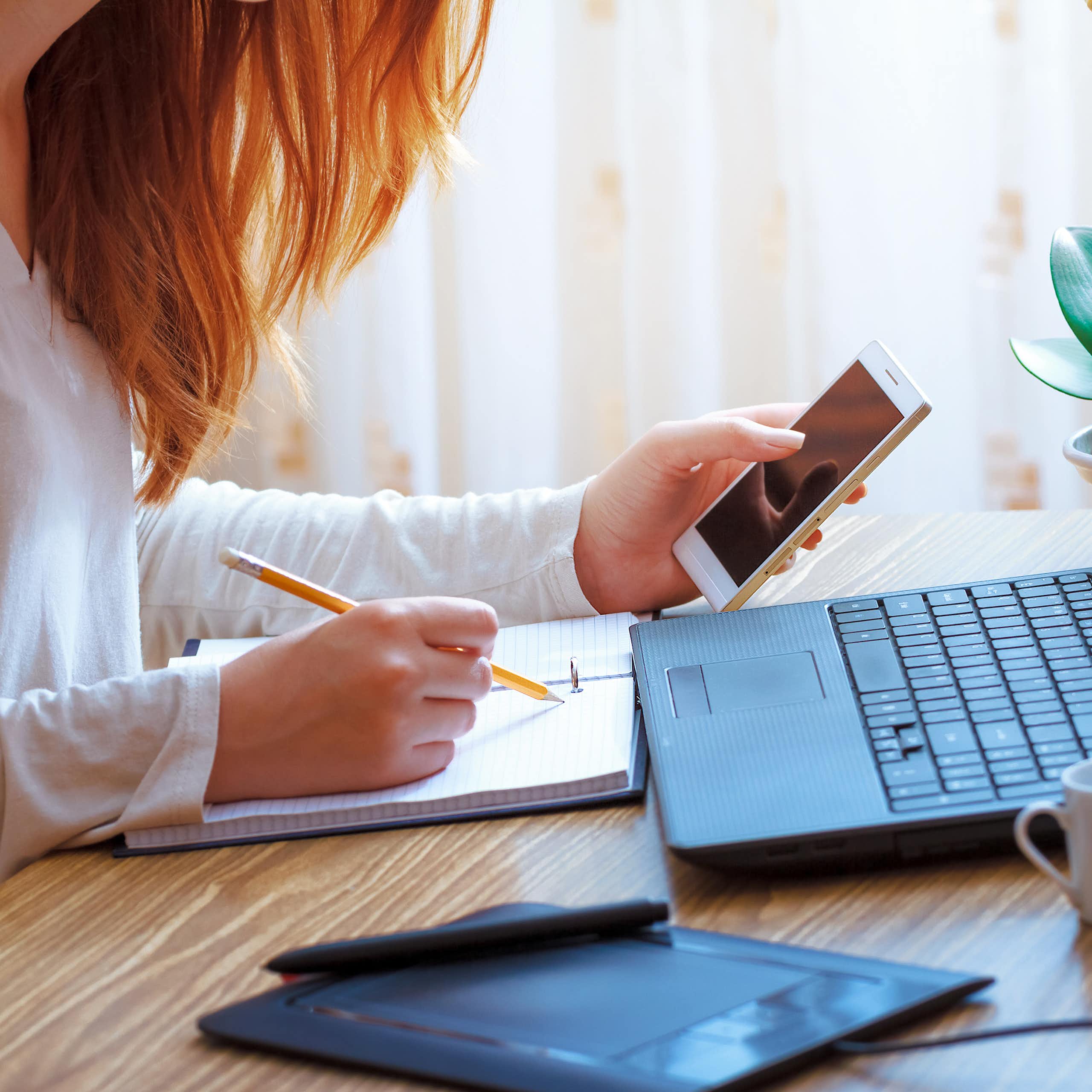 Une jeune femme assise à un bureau, devant un ordinateur portable, consulte son téléphone et écrit en même-temps dans un carnet.