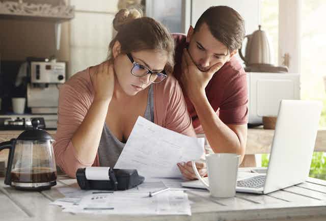 A young couple, looking confused, pores over financial papers at the kitchen counter, looking at a laptop computer