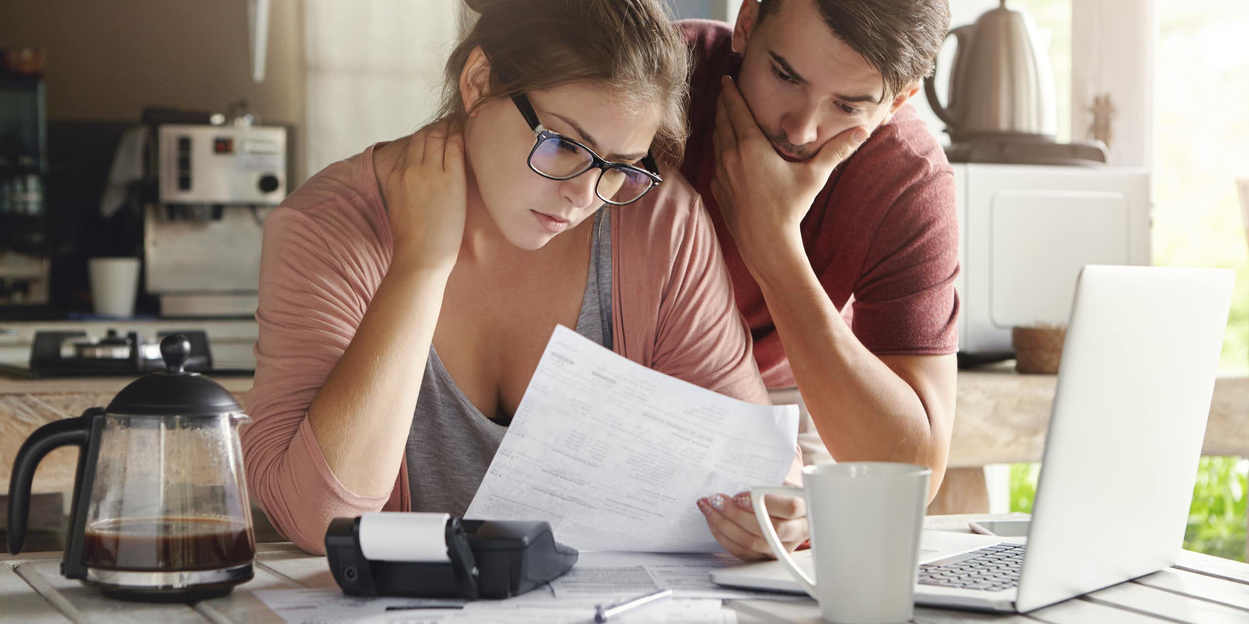 A young couple, looking confused, pores over financial papers at the kitchen counter, looking at a laptop computer
