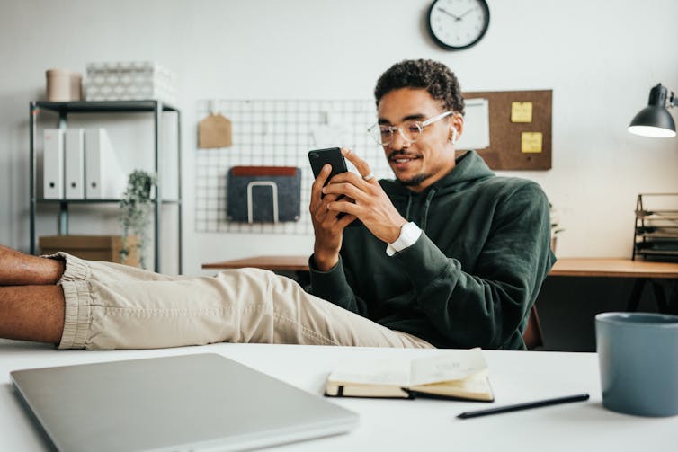 A young black man sits at a home office desk with his feet up, looking at a mobile phone