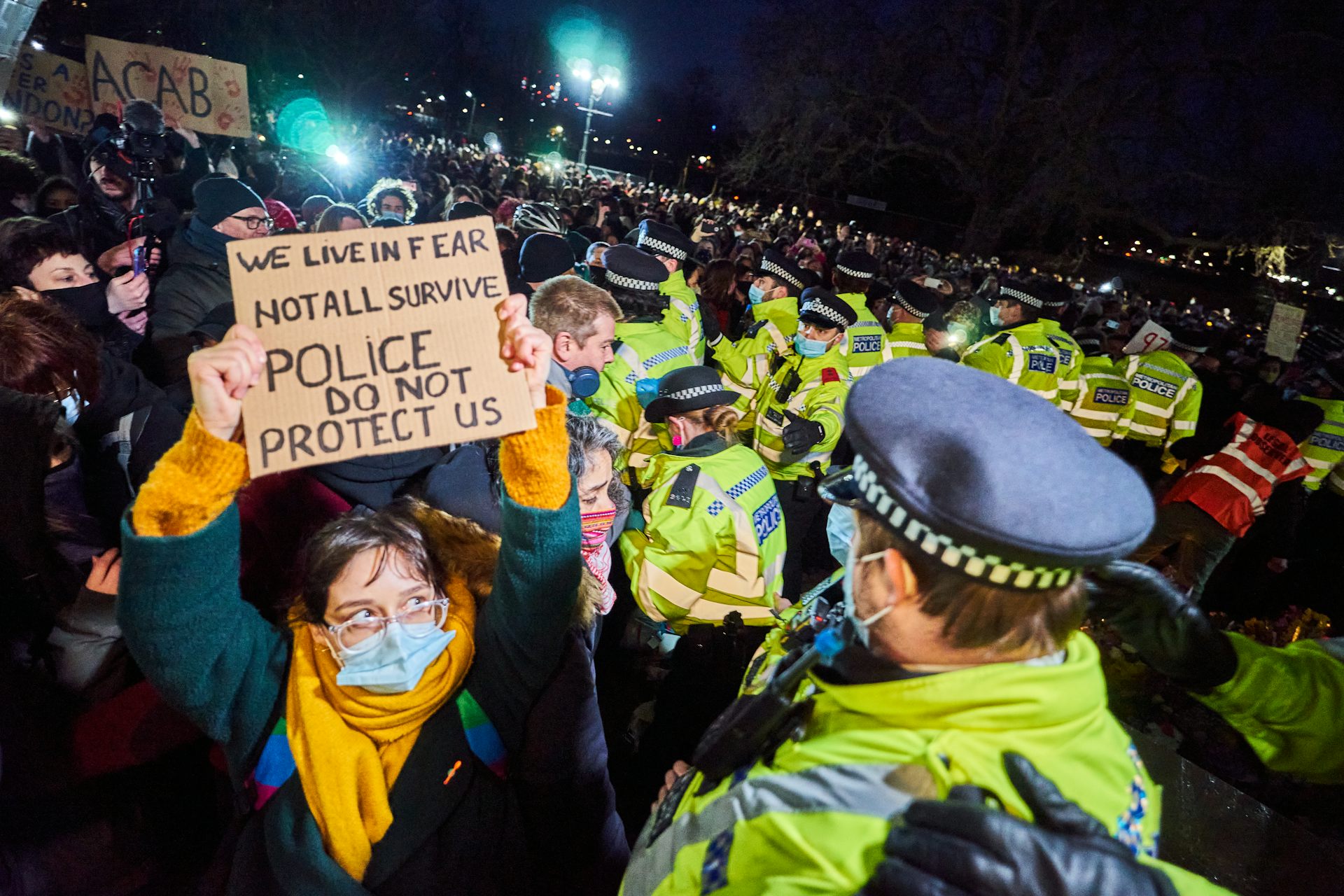 A woman in a face mask holds a handmade cardboard sign reading 'we live in fear, not all survive, police do not protect us'. She is face to face with a police officer, and dozens of other officers and protesters are behind her.