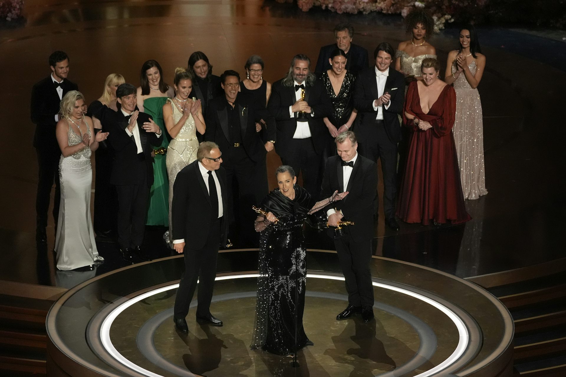 People seen standing on a stage dressed in formal clothing, one holding a golden statue.