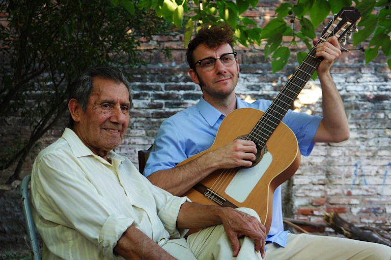 Un hombre joven con una guitarra y un señor mayor miran fuera de campo sonriendo.