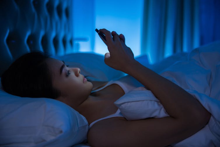 Young Asian woman lying in bed illuminated by the glow of her phone