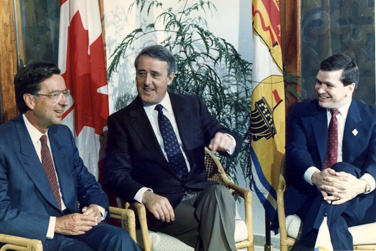 Three men in suits sit and talk in front of a flag.