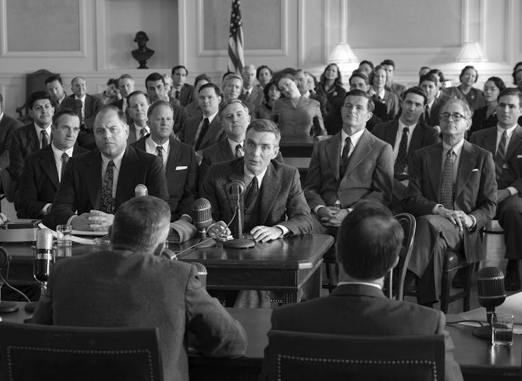 Man in a suit sitting at a long table with a microphone surrounded by other men in suits.