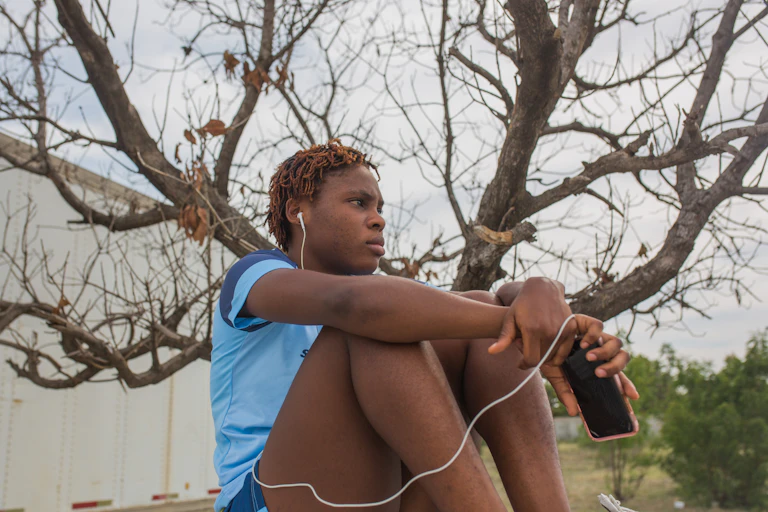 Young Black woman with short hair sits with her arm resting on her knees and listens to music using headphones.