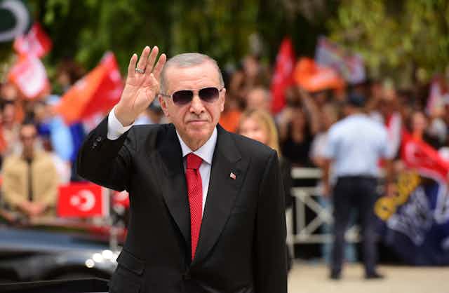 A man in a dark suit and red tie wearing sunglasses waving