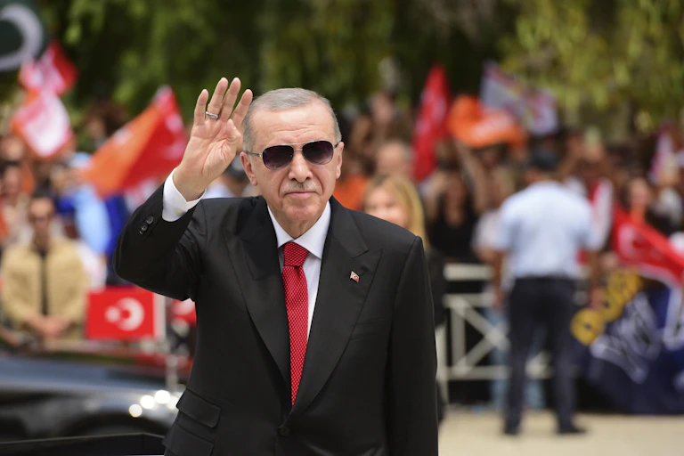 A man in a dark suit and red tie wearing sunglasses waving