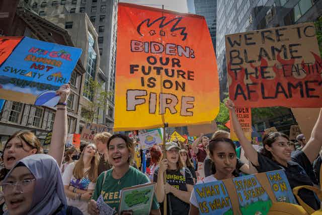 A crowd of young people wear t-shirts and holds signs that say things like 'Biden: our future is on fire' and 'We're to blame, the earth aflame'