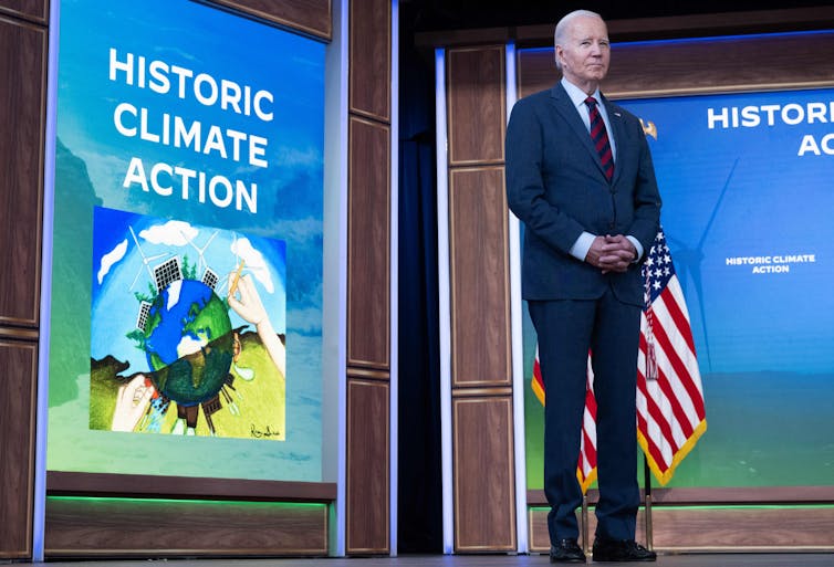 Joe Biden wears a blue suit and stands on a stage in front of a screen that says 'historic climate action.'