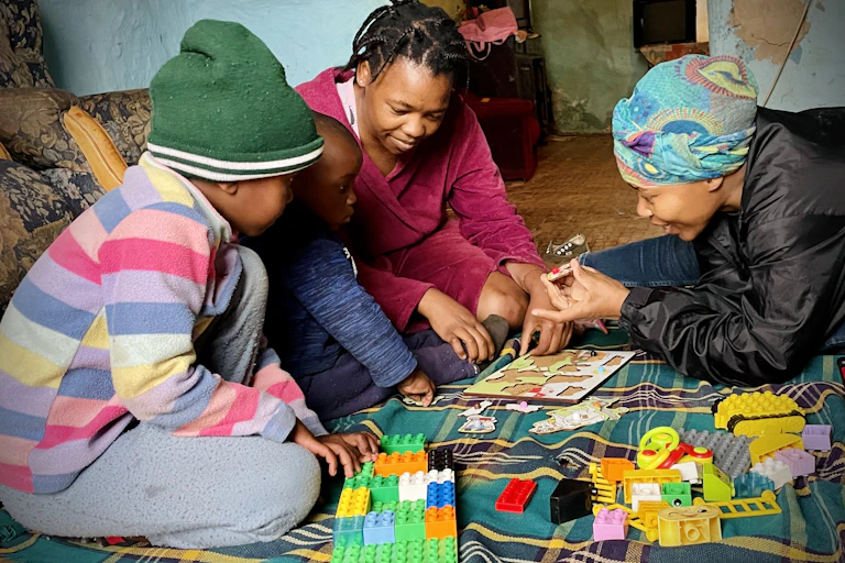 Two adult women play with puzzles, showing pieces to two small children who are not facing the camera
