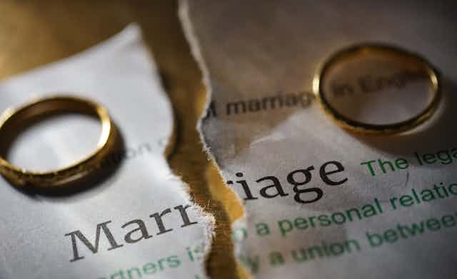 A dictionary page showing the definition of the word "marriage" is torn in two. Two wedding rings are shown resting on the page.
