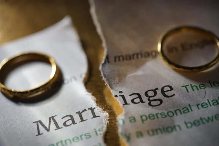 A dictionary page showing the definition of the word "marriage" is torn in two. Two wedding rings are shown resting on the page.