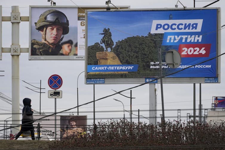 A woman walks past a billboard with Russian words on and another will a soldier's head in a helmet depicted.