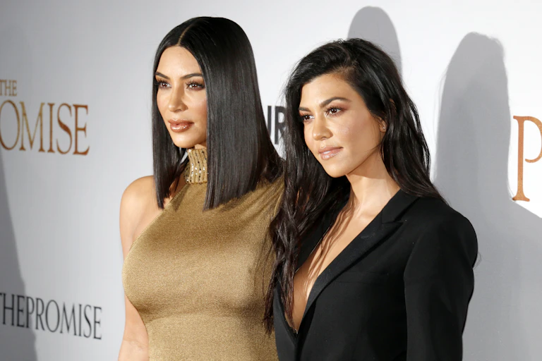 Kim Kardashian con un vestido dorado sin mangas posa junto a Kourtney Kardashian en un evento.