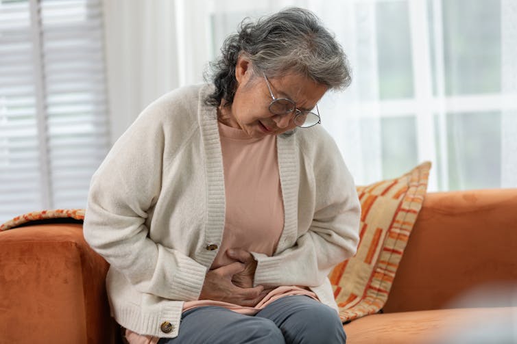 older woman sits on couch and shows pained expression, clutches lower stomach