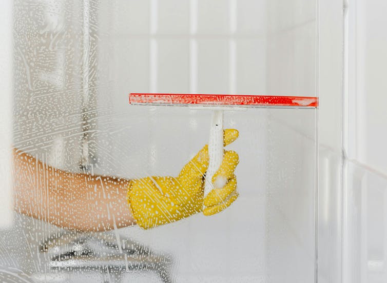 A hand in a yellow glove cleaning the inside of a shower screen with a squeegee.