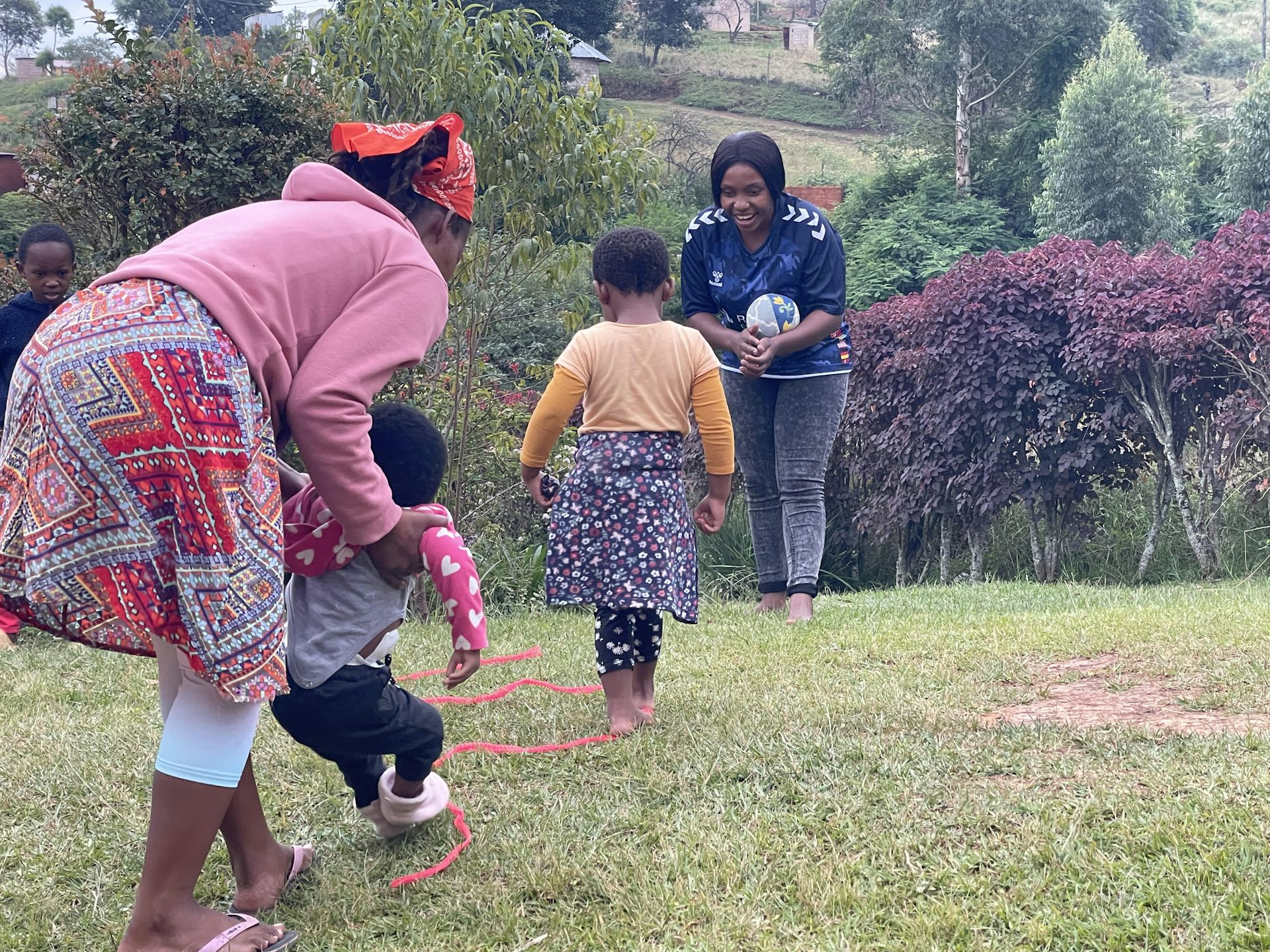 One adult woman holds up a small child while another child walks towards an adult woman holding a ball