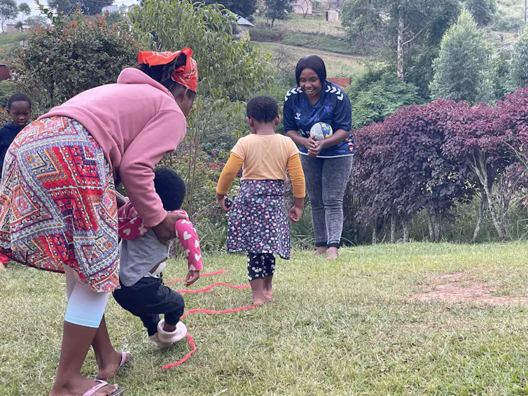 One adult woman holds up a small child while another child walks towards an adult woman holding a ball